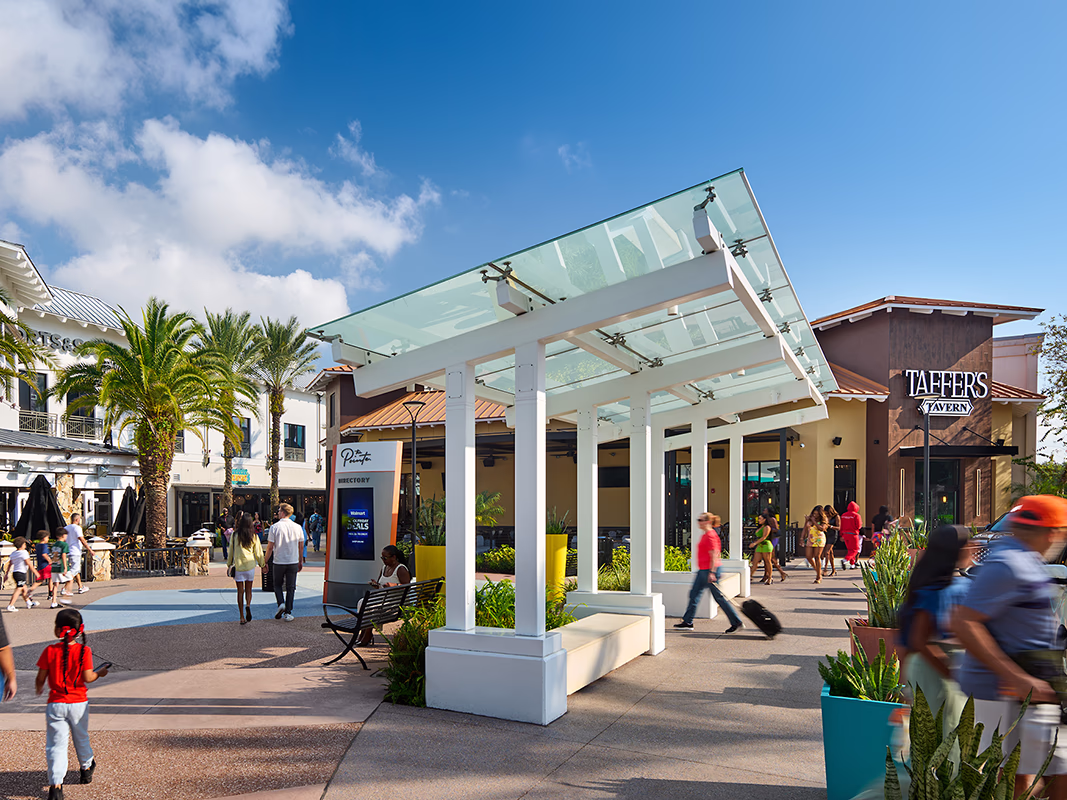 Visitors walking through Pointe Orlando's open-air plaza on a sunny day beneath a modern glass and white steel canopy structure, with a digital directory kiosk, palm trees, and storefronts visible in the background.