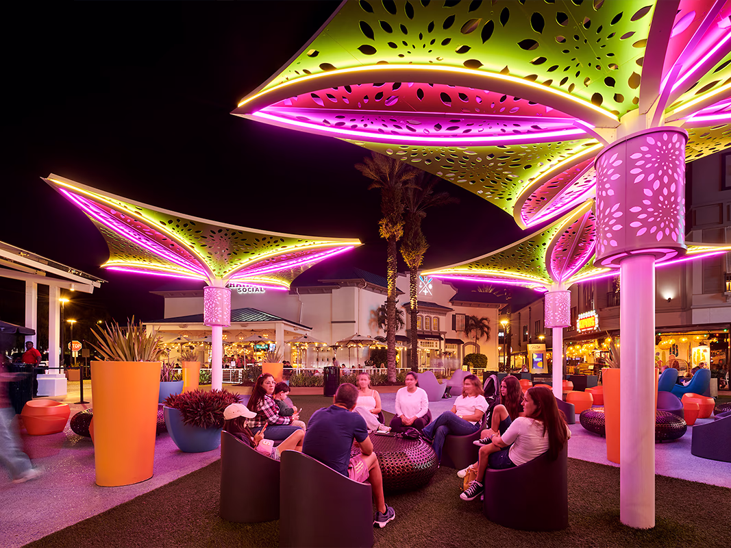 Groups of people socializing on curved lounge seating beneath large flower-shaped canopy structures with glowing green and pink neon LED lighting at Pointe Orlando's outdoor plaza at night.