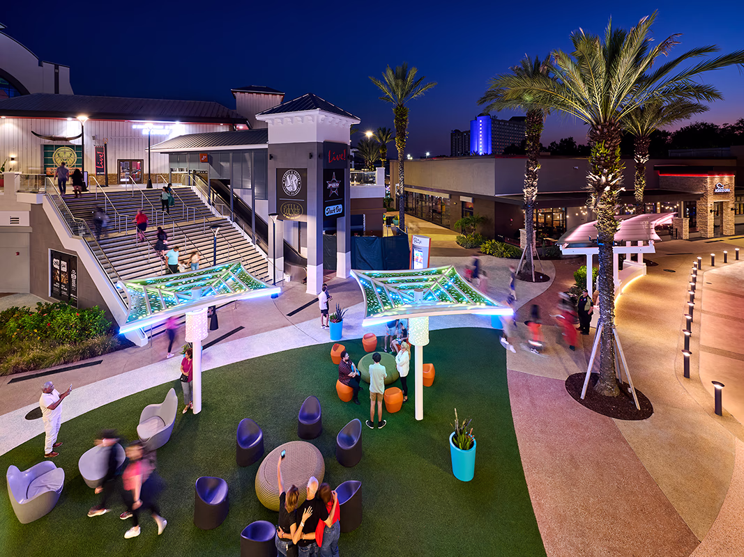 Aerial view of Pointe Orlando's outdoor plaza at dusk, with people mingling around colorful lounge seating and a glowing LED canopy structure, surrounded by palm trees, retail storefronts, and a grand staircase leading to an upper level.