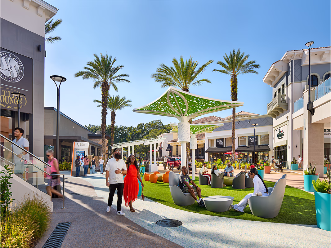 Visitors walking and relaxing in modern pod-style lounge chairs on a turf area at Pointe Orlando's outdoor plaza during the day, with a sculptural green perforated canopy structure, tall palm trees, and retail storefronts lining the walkway under a bright blue sky.