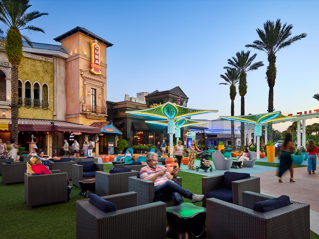 People relaxing in wicker lounge chairs and colorful seating at Pointe Orlando's outdoor plaza at dusk, with glowing teal LED canopy structures, lush turf, and palm trees set against a clear evening sky.