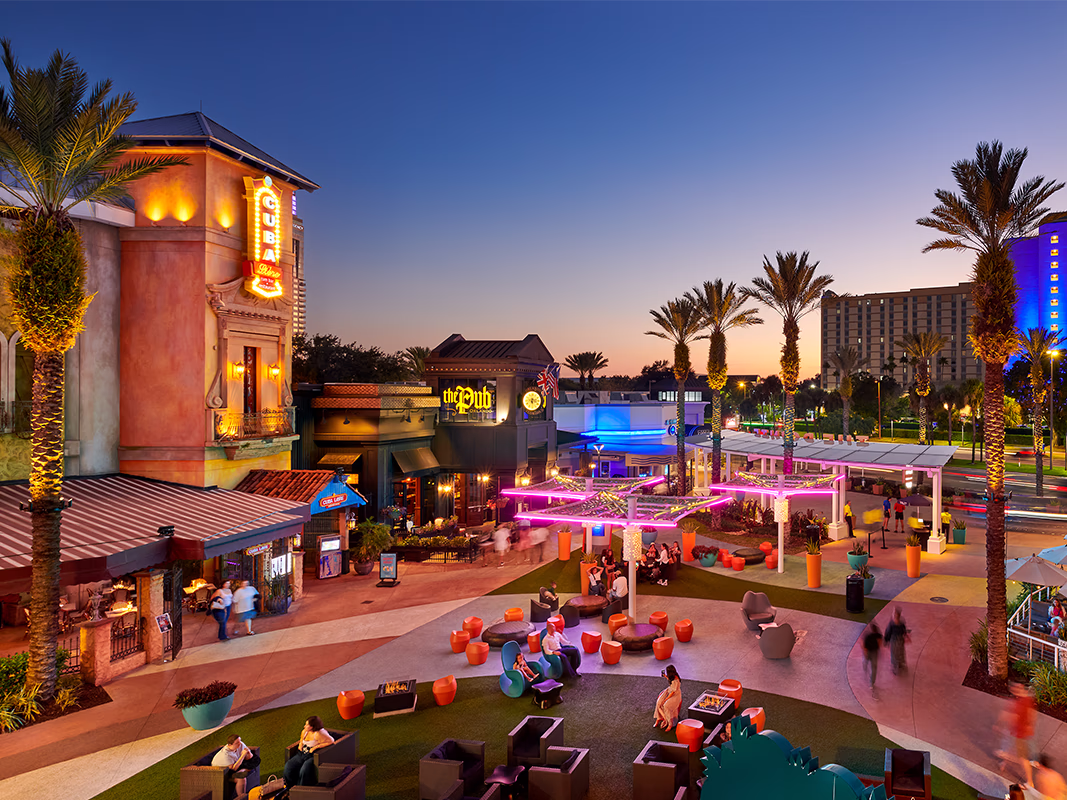 Elevated view of The Pointe Orlando's central plaza at dusk, showing lounge seating, illuminated shade structures, palm trees, and multiple restaurant tenants including Cuba Libre and The Pub, with the International Drive corridor visible in the background.