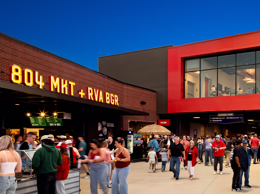 Fans walk through the evening concourse past illuminated '804 MKT + RVA BGR' food signage, a tiki-style umbrella bar, and section wayfinding signs.