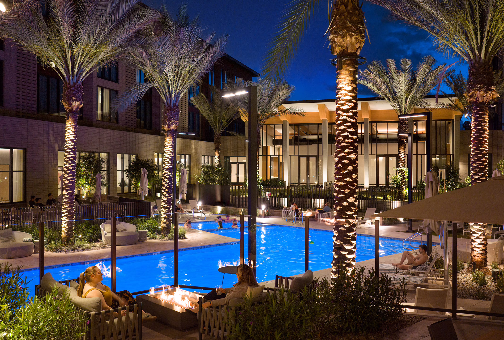 Guests relax around a fire pit lounge at the Norterra hotel's courtyard pool in the evening, with uplit palm trees and the shared amenity building in the background