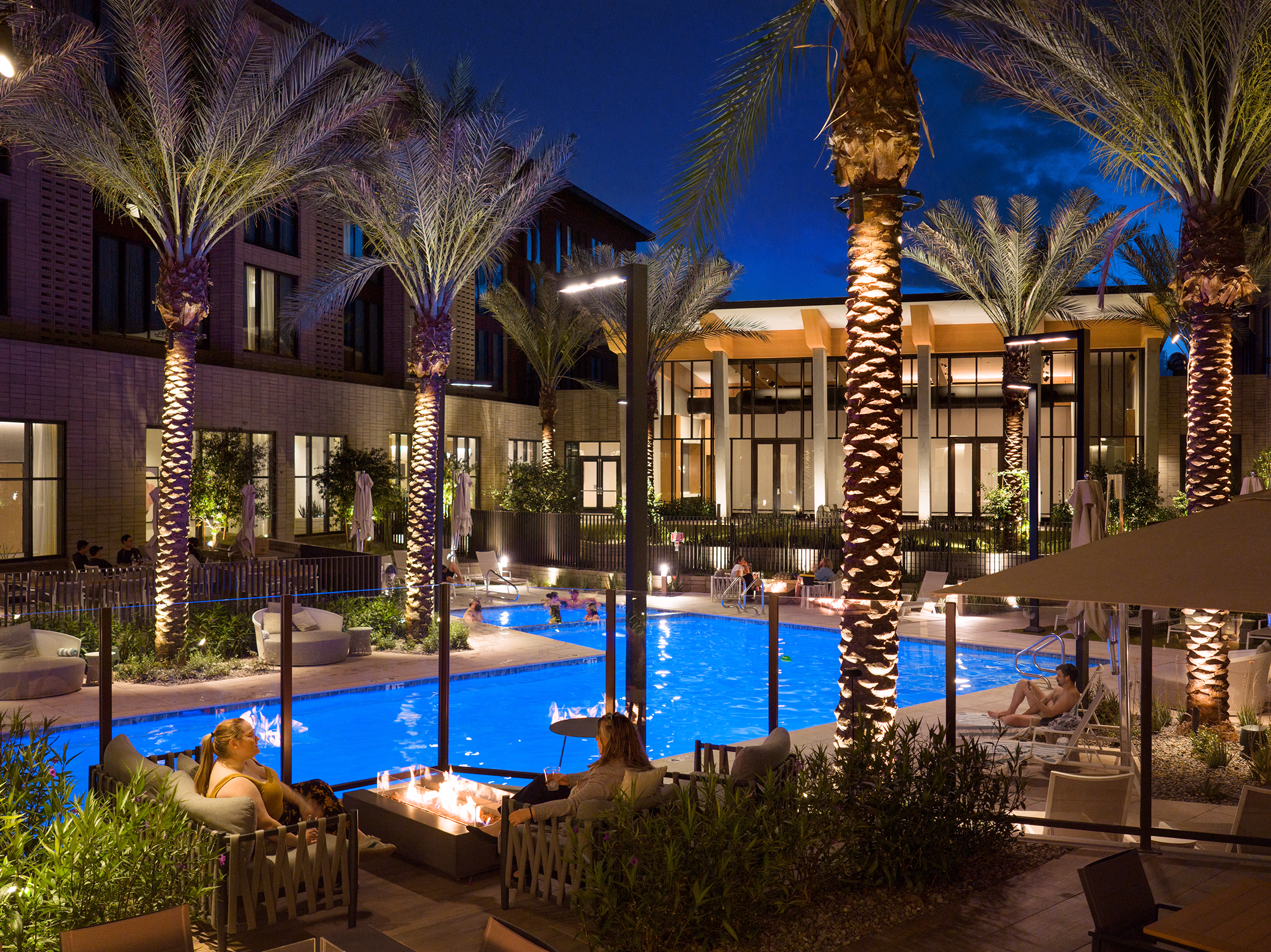 Guests relax around a fire pit lounge at the Norterra hotel's courtyard pool in the evening, with uplit palm trees and the shared amenity building in the background
