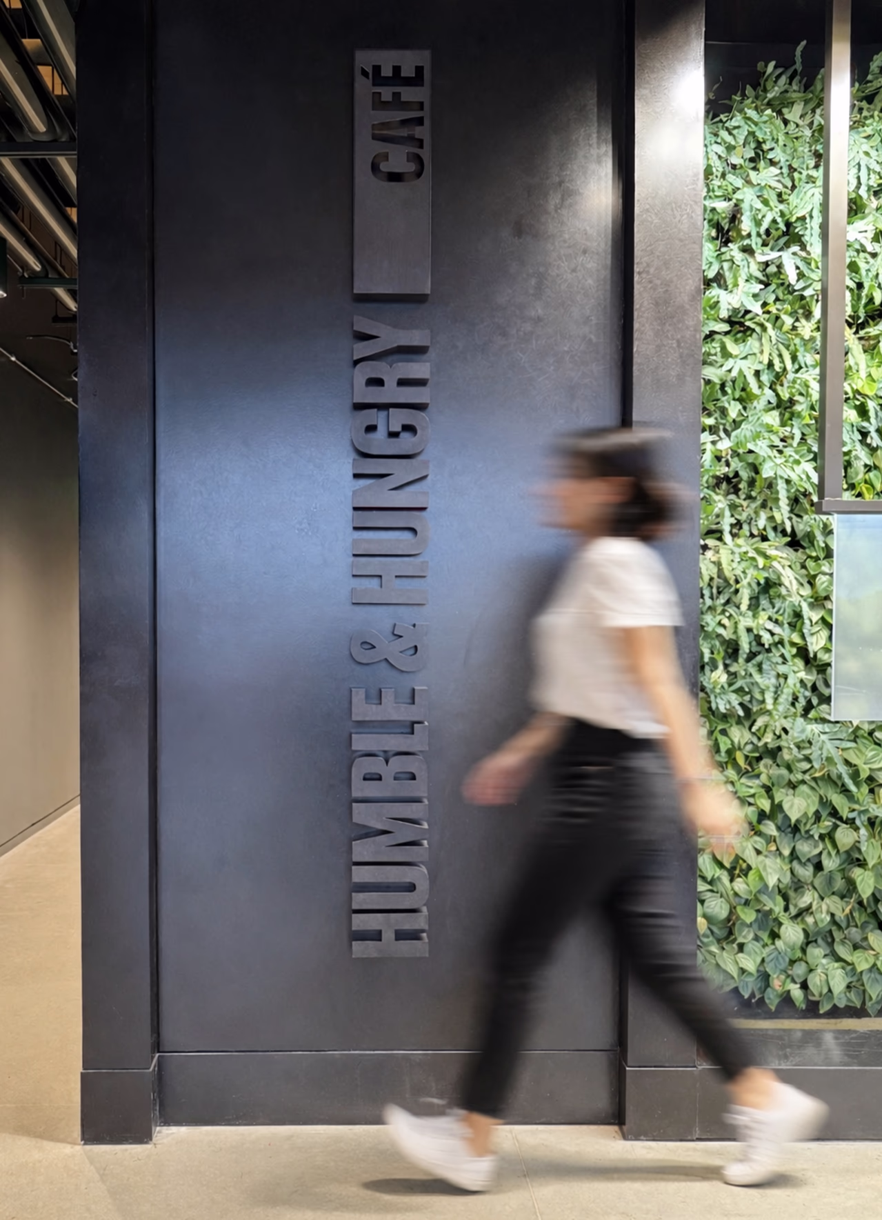 Close-up of dimensional 'Humble & Hungry Café' entrance signage at Under Armour headquarters, mounted on a dark wall beside a living green wall, with a person walking past.