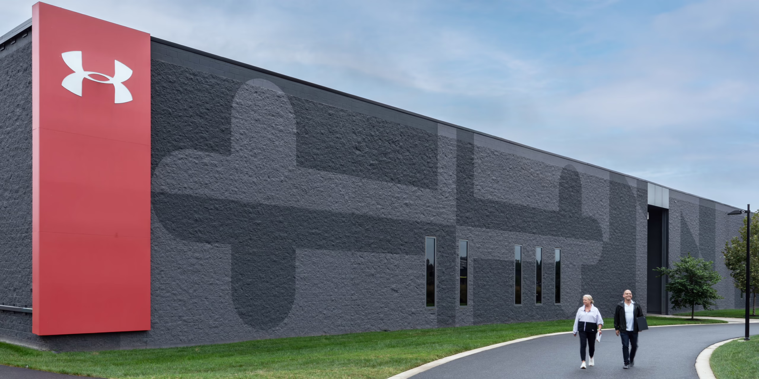 Exterior view of a dark facade campus building at Under Armour headquarters showing a subtle tonal wordmark pattern across the brick wall, with two people walking along the entry path.