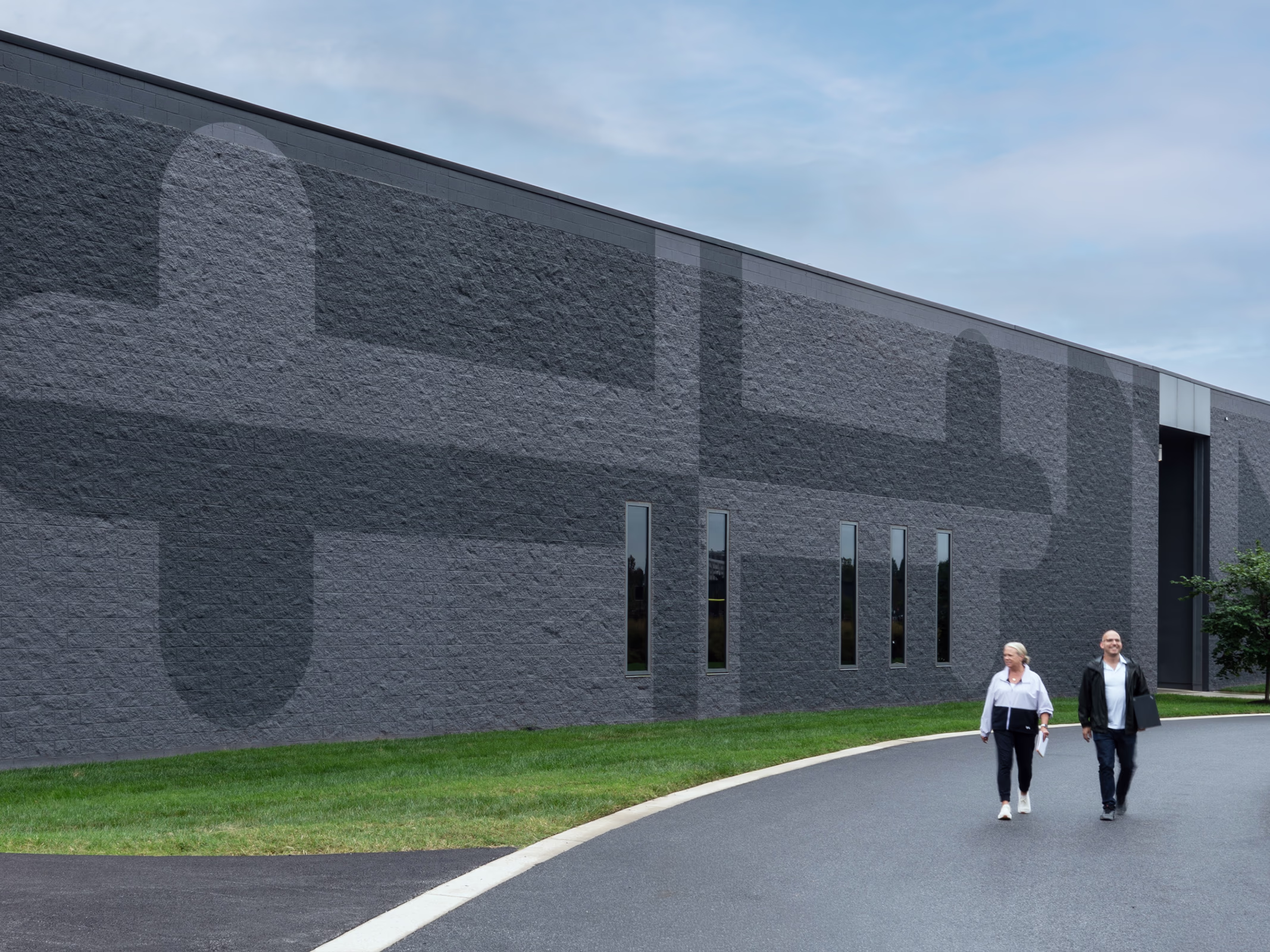 Exterior view of a dark facade campus building at Under Armour headquarters showing a subtle tonal wordmark pattern across the brick wall, with two people walking along the entry path.