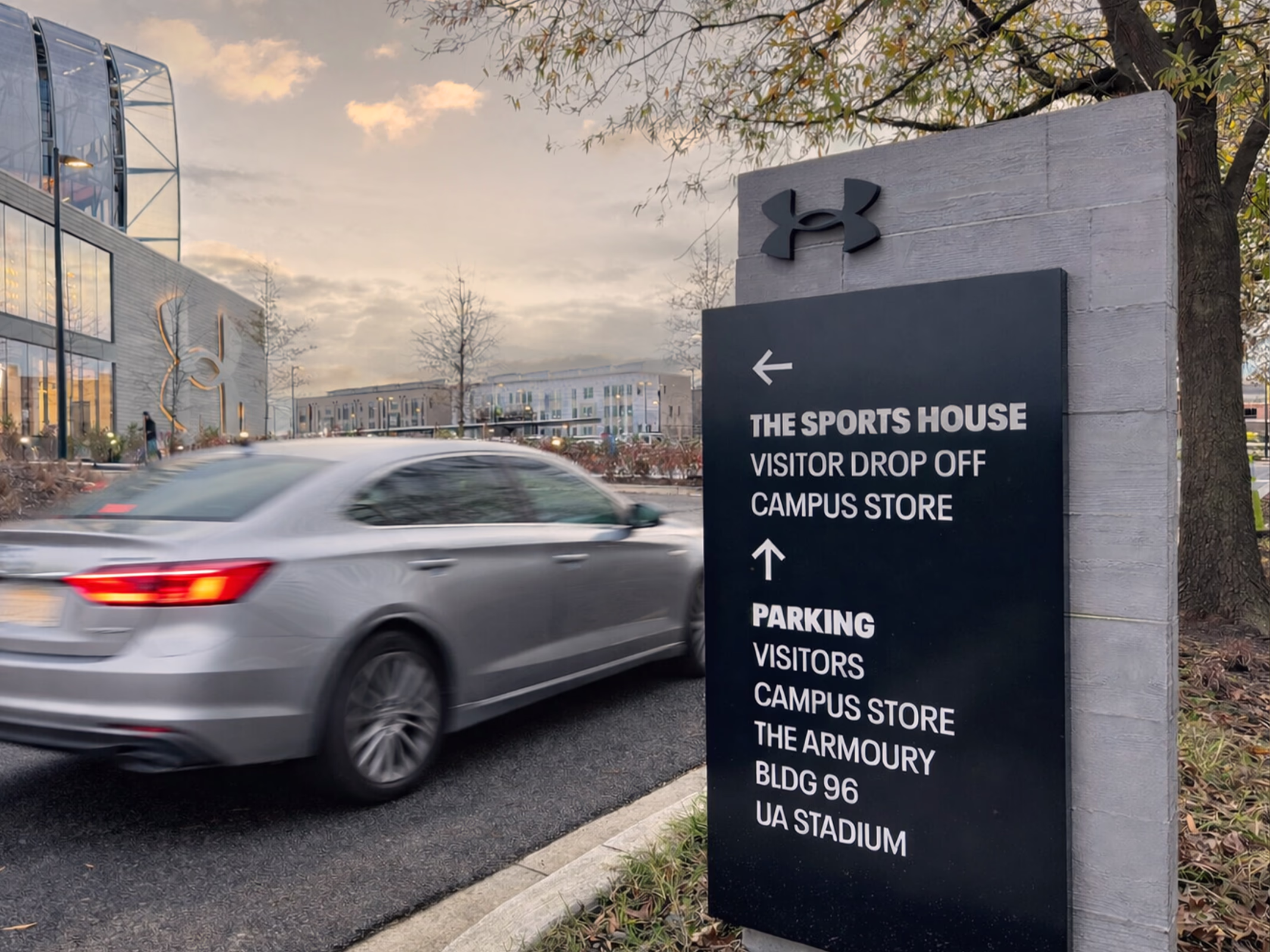 Vehicular directional sign at Under Armour headquarters campus directing visitors to The Sports House, visitor drop off, campus store, and parking, with a car passing by and the headquarters building in the background.