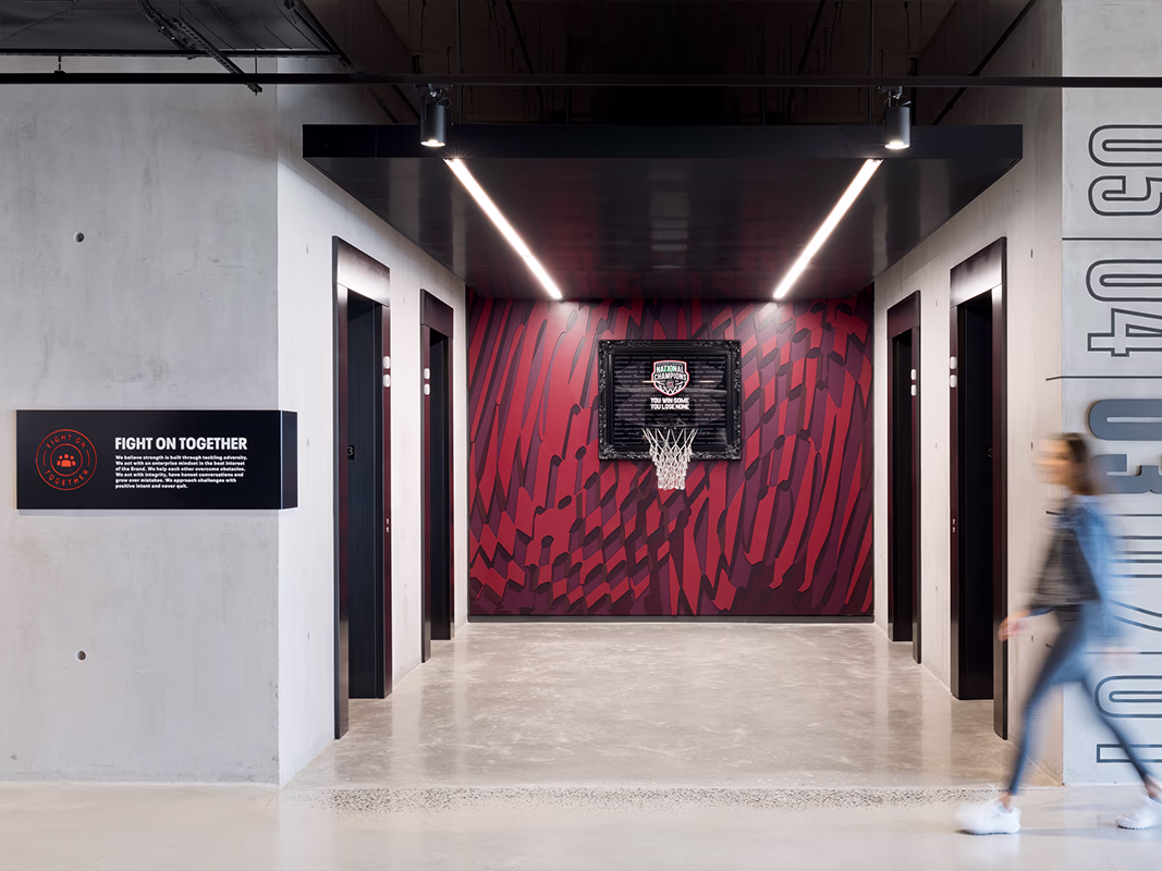 Elevator lobby at Under Armour headquarters featuring a bold red patterned wall graphic with a framed 'National Champions' basketball hoop display, a 'Fight on Together' brand values sign, and floor-number graphics on the concrete wall, with a person walking through.