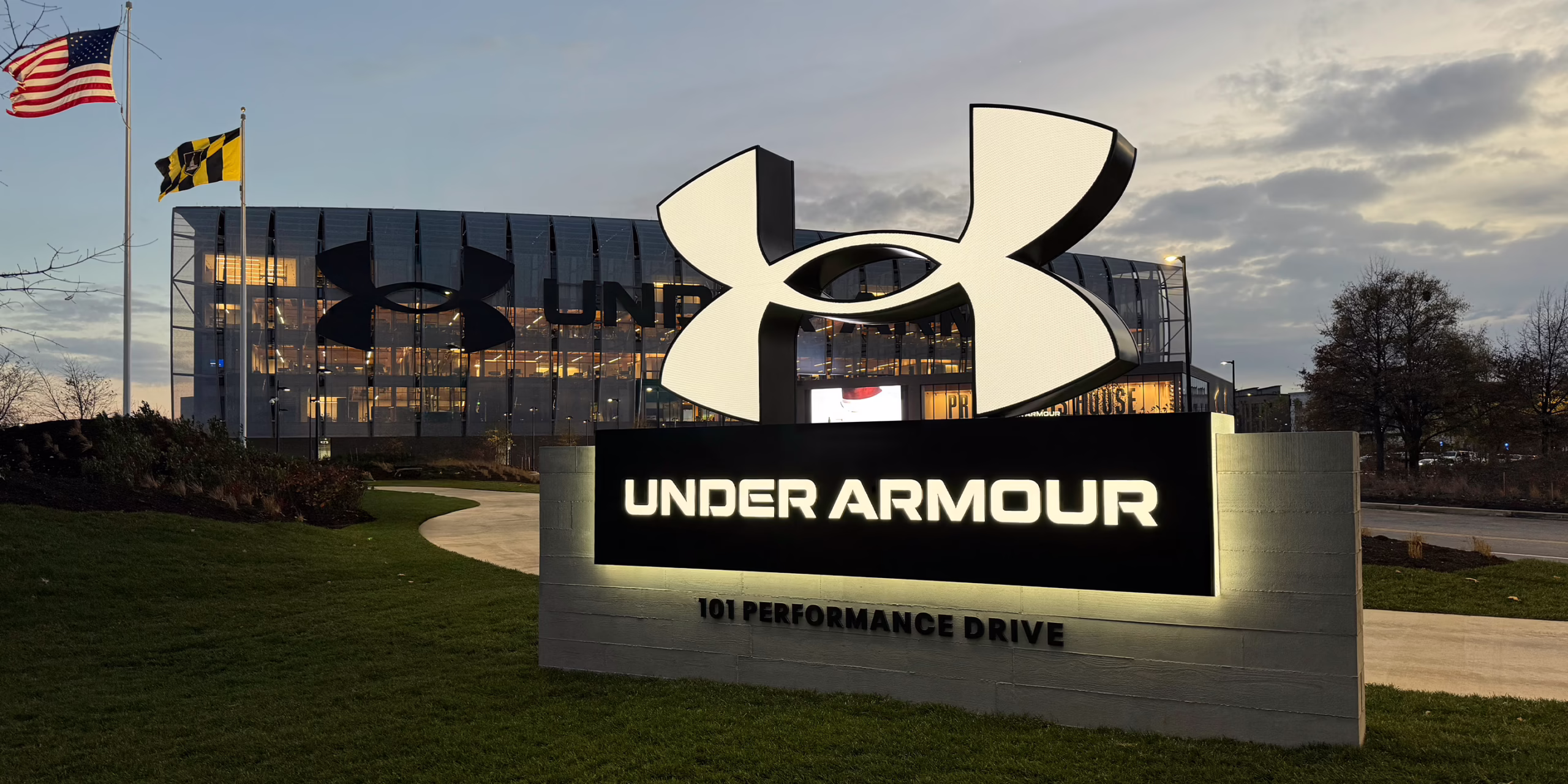 Illuminated exterior monument sign reading 'Under Armour, 101 Performance Drive,' with a large sculptural logo above it and the glass headquarters facade lit up at dusk in the background.