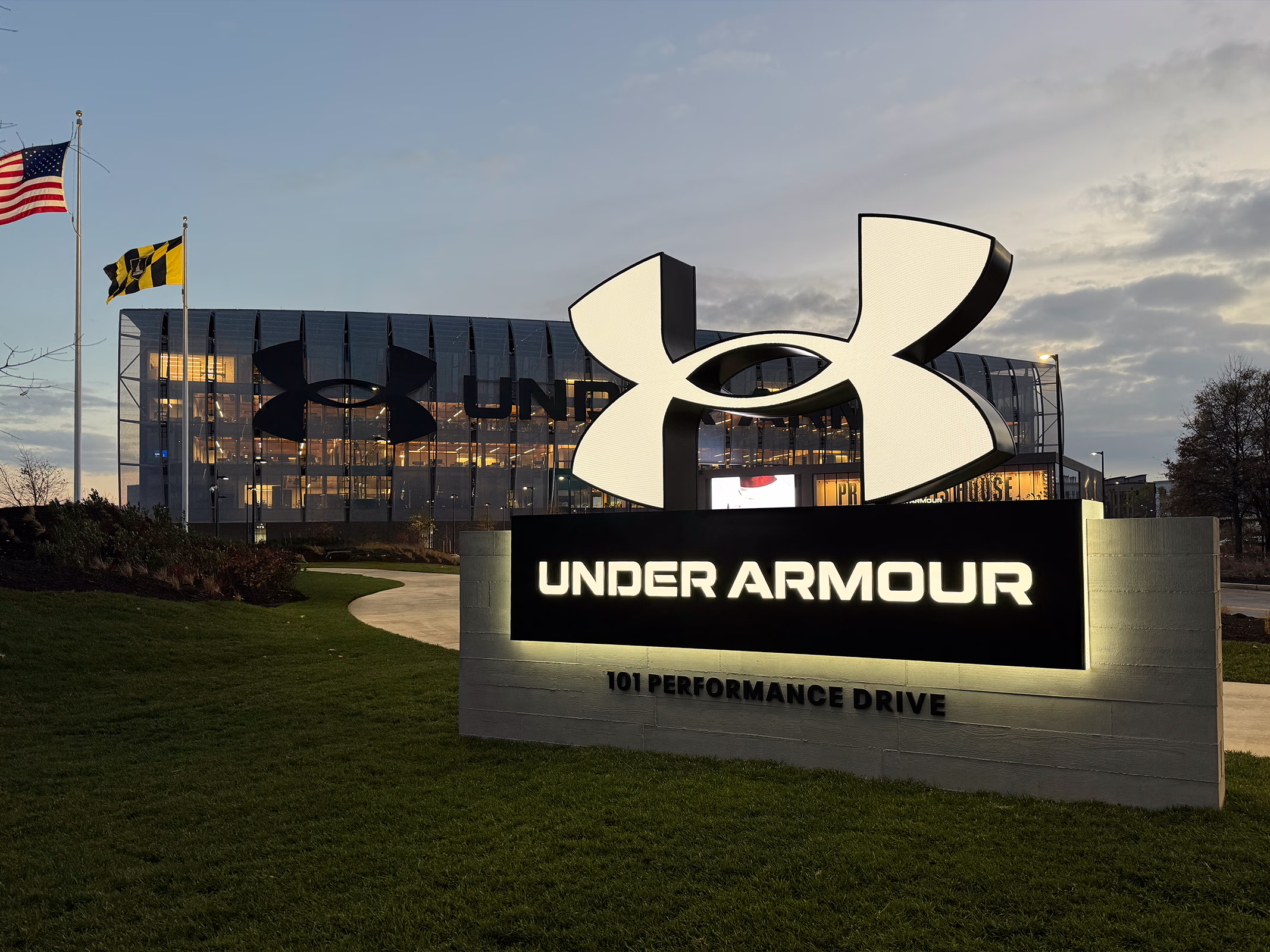 Illuminated exterior monument sign reading 'Under Armour, 101 Performance Drive,' with a large sculptural logo above it and the glass headquarters facade lit up at dusk in the background.