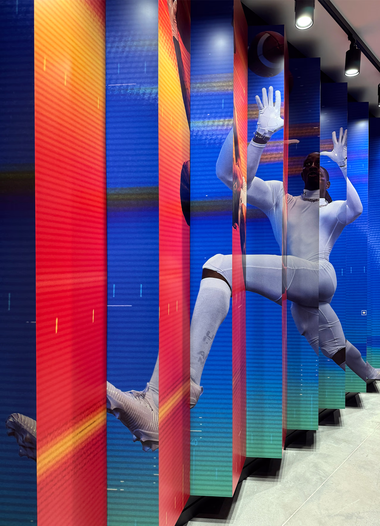 Close-up of a fin-panel wall installation at Under Armour headquarters displaying dynamic athlete photography in a football-inspired motion catch, with a colorful gradient background.