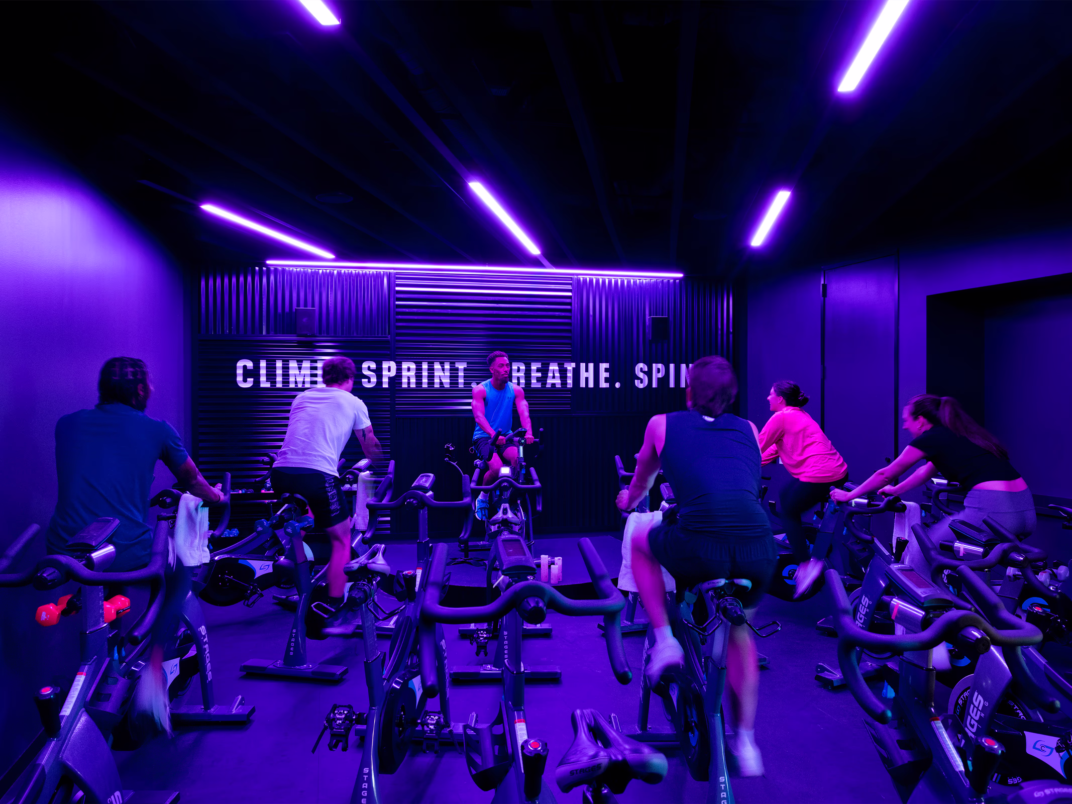 Indoor cycling studio at Under Armour headquarters lit in purple, with employees on spin bikes facing a wall reading 'Climb. Sprint. Breathe. Spin.