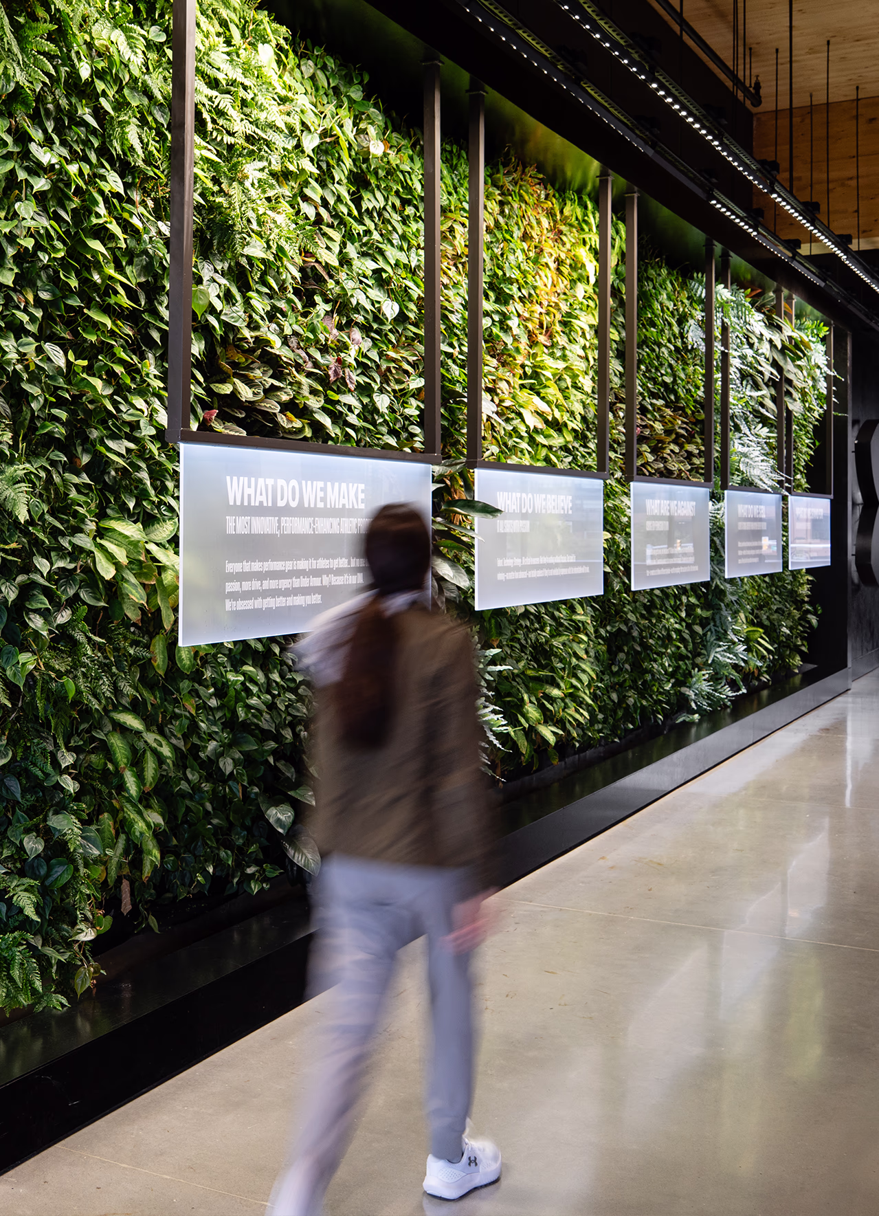 Vertical view of a living green wall at Under Armour headquarters with backlit panels reading 'What Do We Make' and 'What Do We Believe,' as a person walks past in the corridor.