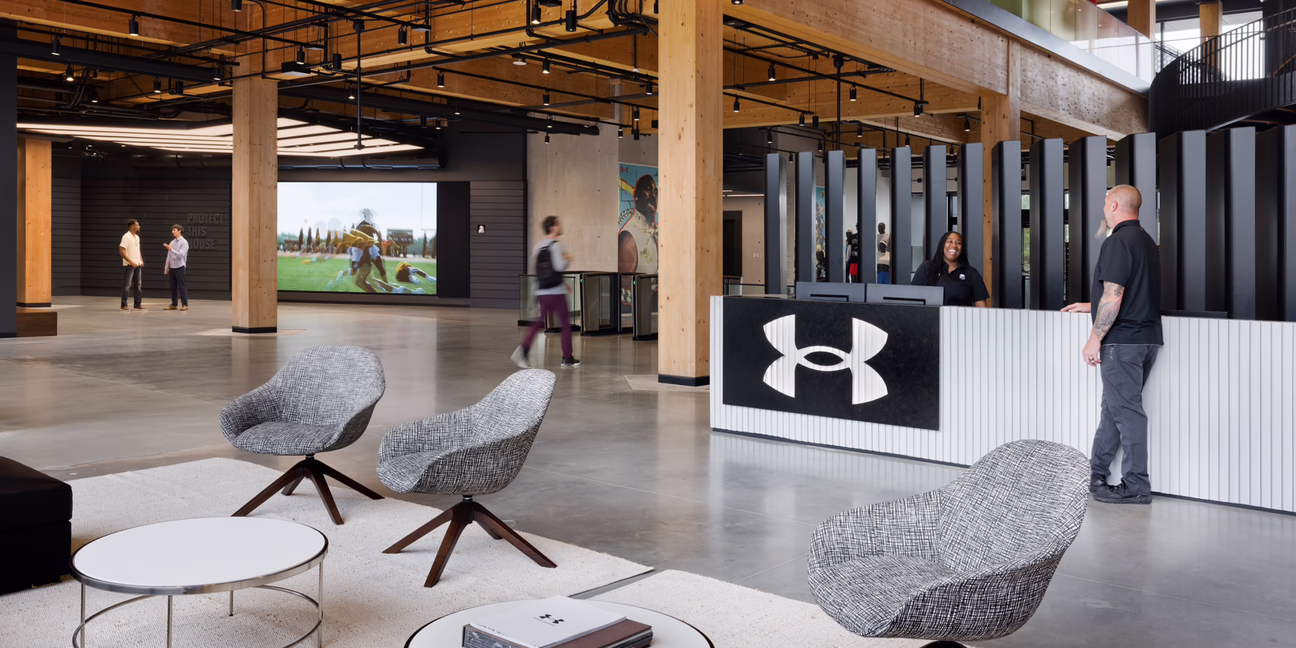 Main reception lobby at Under Armour headquarters featuring a large illuminated logo panel at the front desk, lounge seating, a large video display showing athletic footage, and wood ceiling beams throughout the open space.