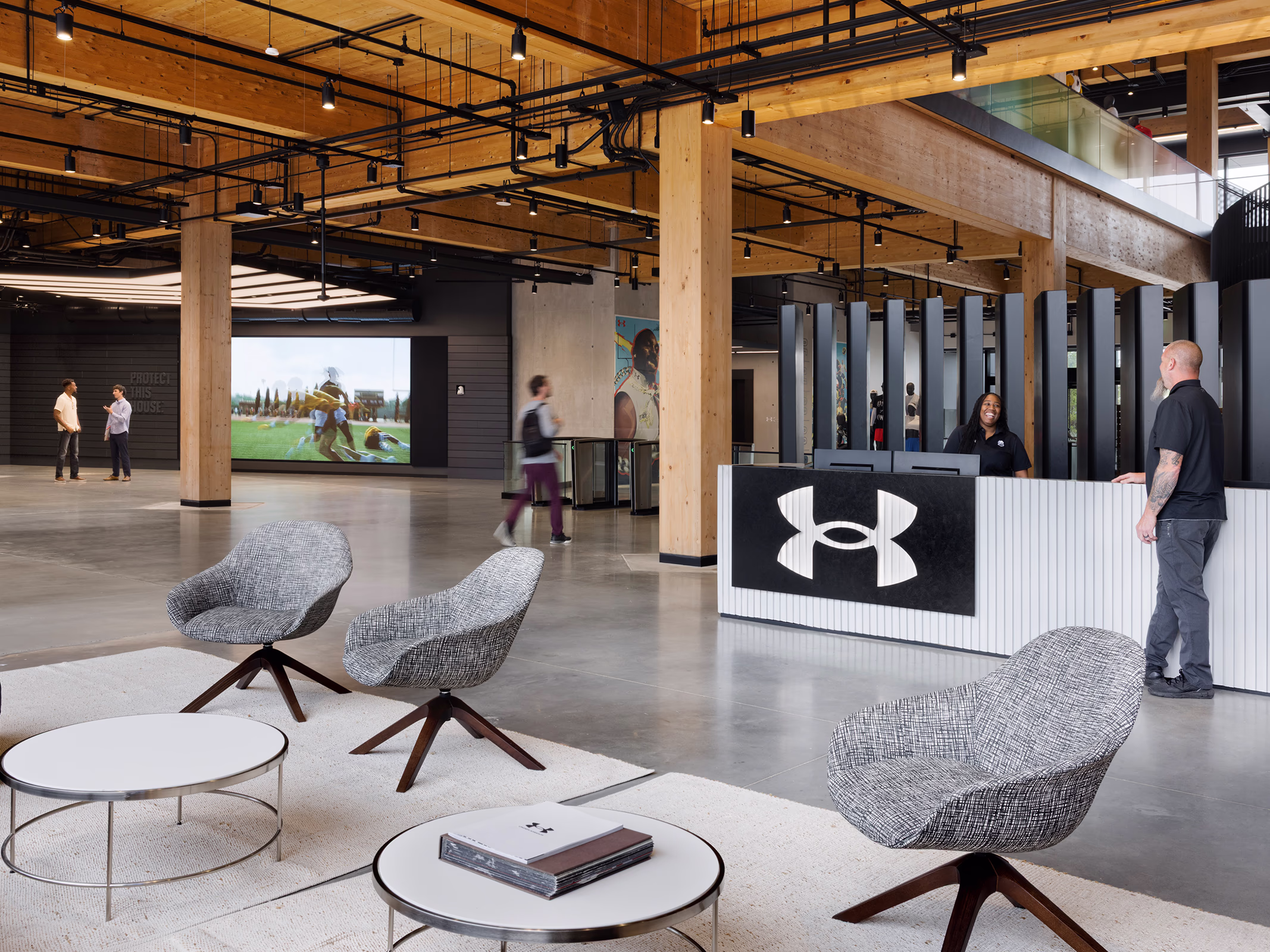 Main reception lobby at Under Armour headquarters featuring a large illuminated logo panel at the front desk, lounge seating, a large video display showing athletic footage, and wood ceiling beams throughout the open space.