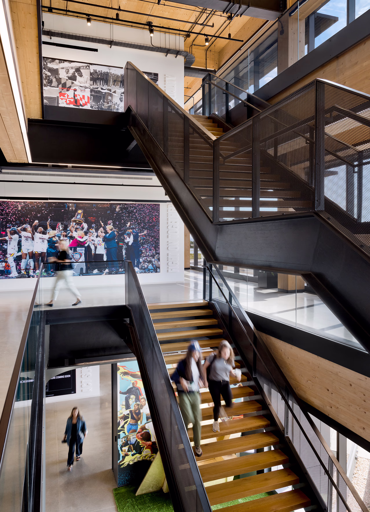 Multi-level steel and wood staircase at Under Armour headquarters with employees moving between floors, framed by large-scale black-and-white historical photography and a championship celebration photo mural on the atrium walls.
