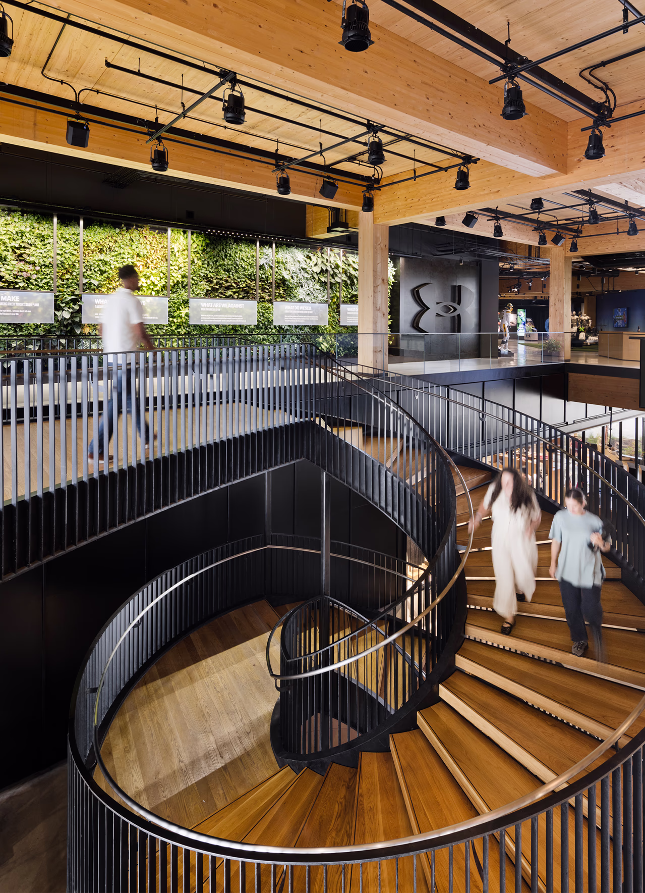 Wood and steel spiral staircase at Under Armour headquarters with employees descending, overlooking a living green wall with brand storytelling panels on the level above.